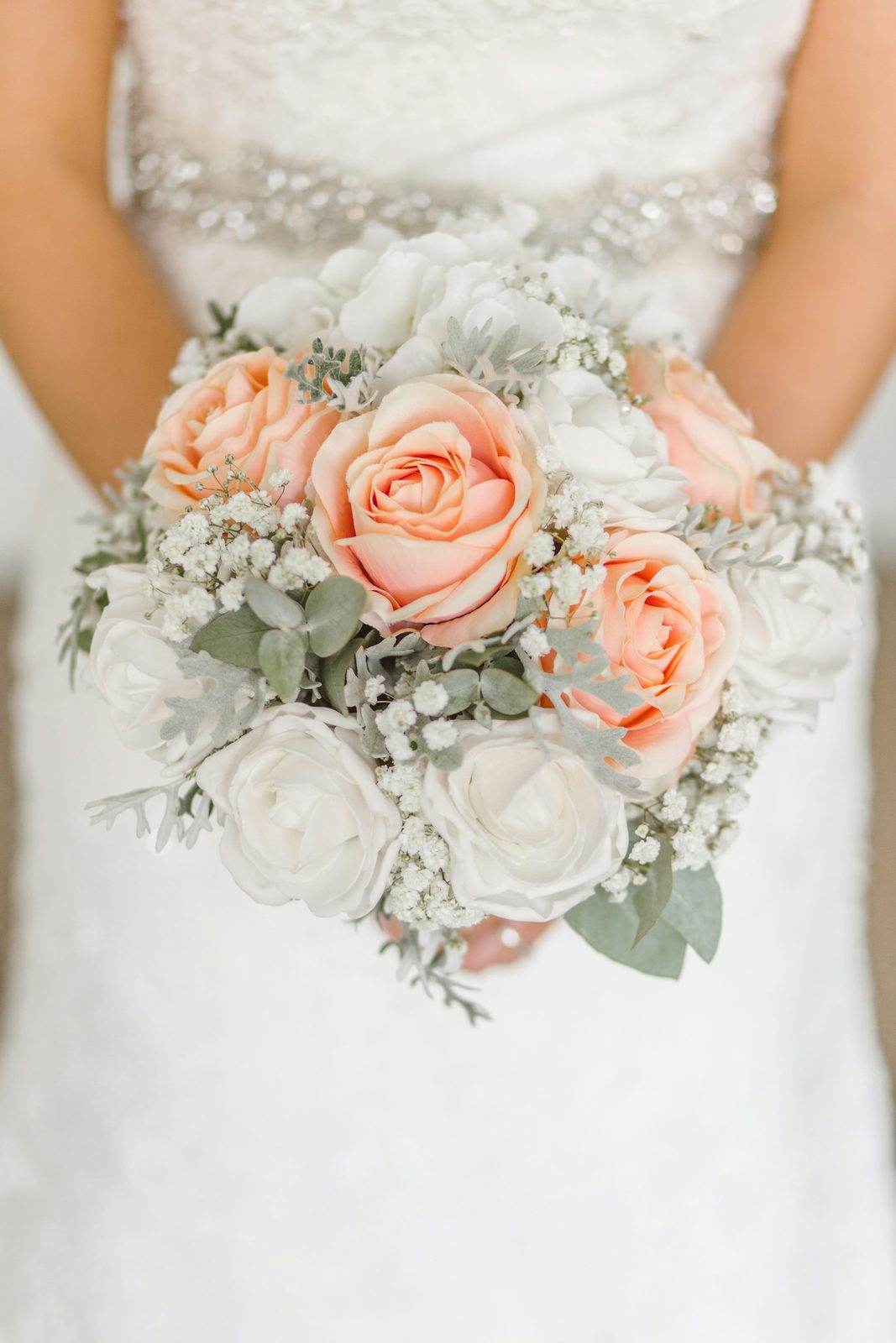 Bride holding a bouquet of peach roses and white blooms against a beaded gown