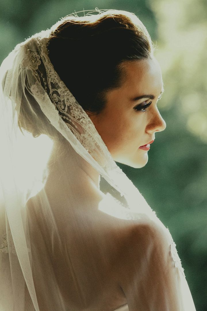 Bride in profile wearing a lace-edged veil over a low updo, greenery behind
