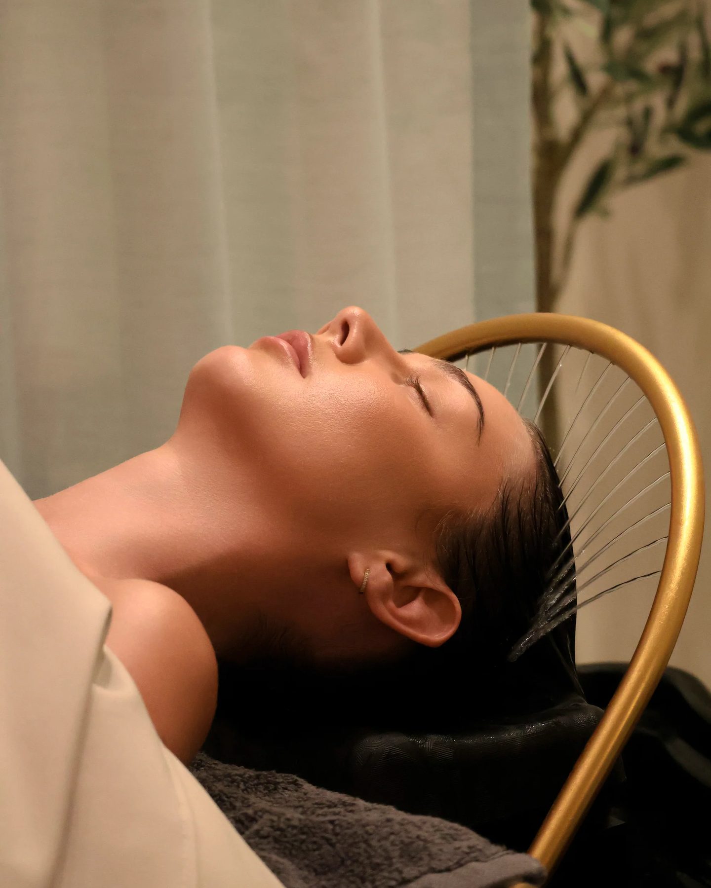 Guest resting with eyes closed as a fine arc of water from a gold head-spa rail rinses her hair